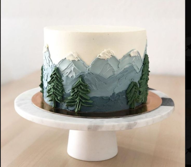 a white and blue cake with trees on it sitting on top of a wooden table