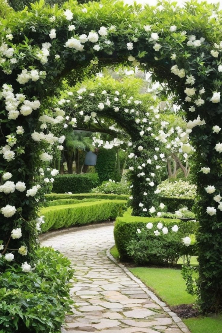 an archway with white flowers on it in the middle of a garden