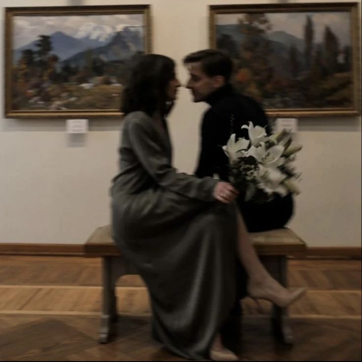 a man and woman sitting next to each other on a bench in front of paintings