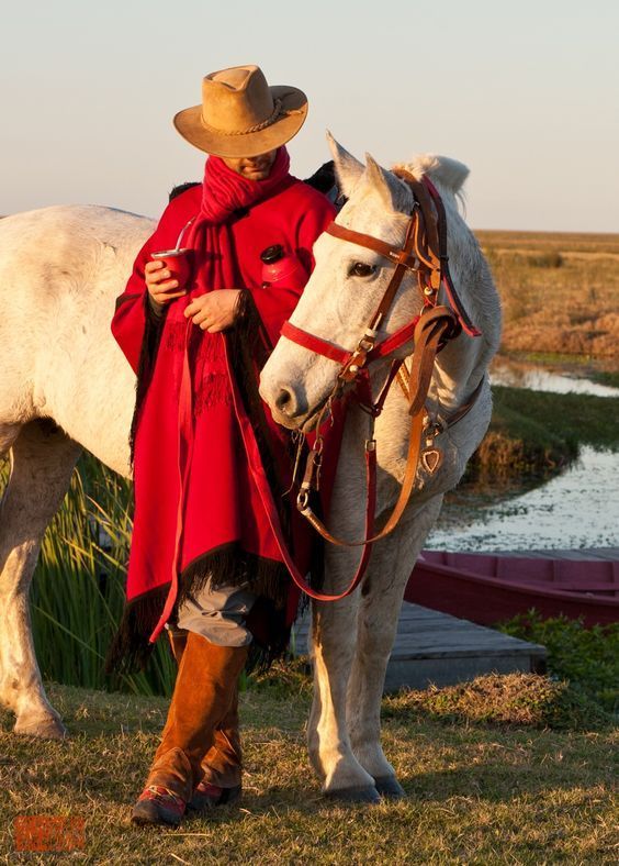 a man in a red coat and cowboy hat standing next to a white horse on the grass