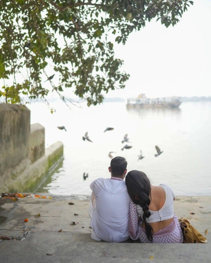 a man and woman sitting on the ground next to water with birds flying around them