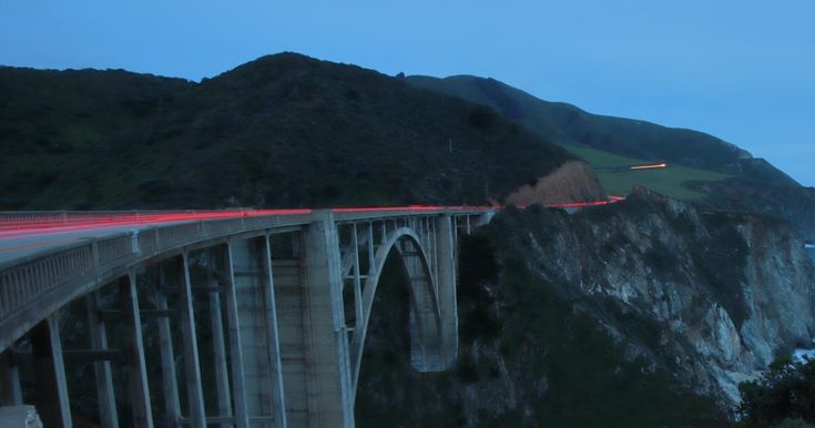 a long bridge over the ocean with cars driving on it at night near mountains and water