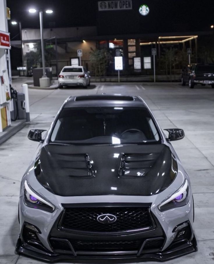 the front end of a gray sports car parked in a parking lot with gas pumps