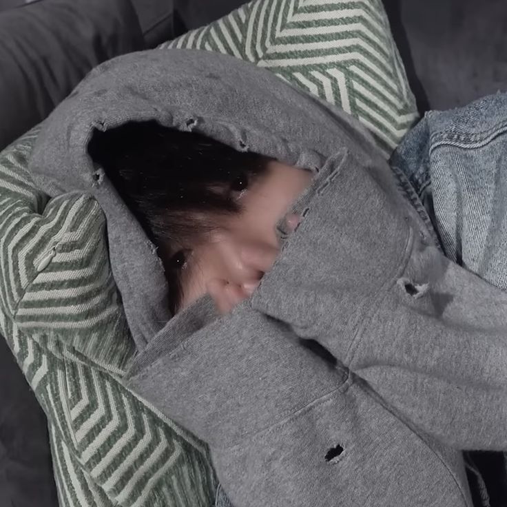 a woman laying on top of a couch covered in a gray hoodie with her eyes closed