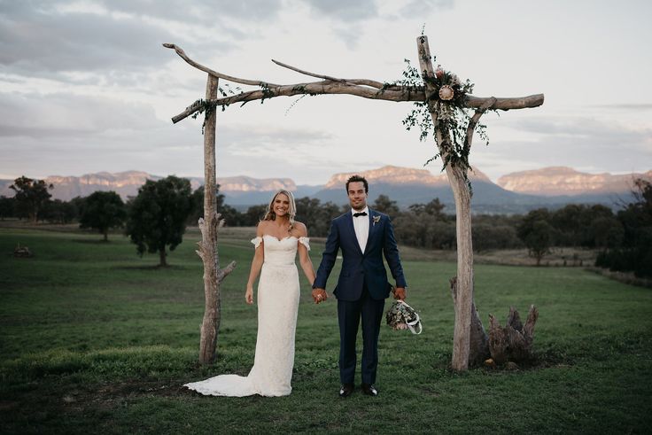 a bride and groom holding hands under an arch made out of branches with mountains in the background