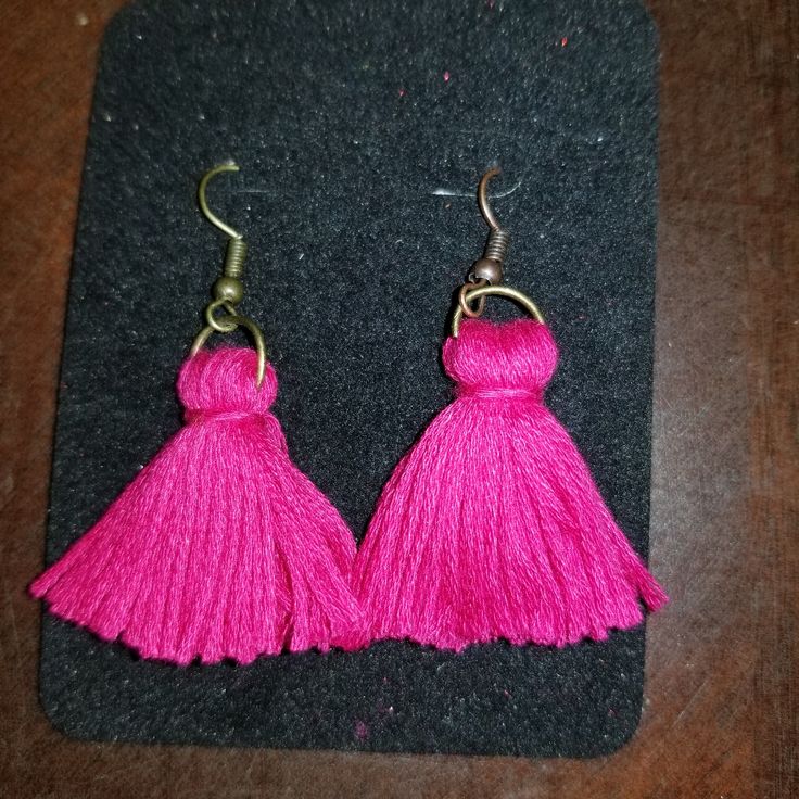 two pink tasselled earrings on top of a black piece of cloth next to a pair of metal hooks