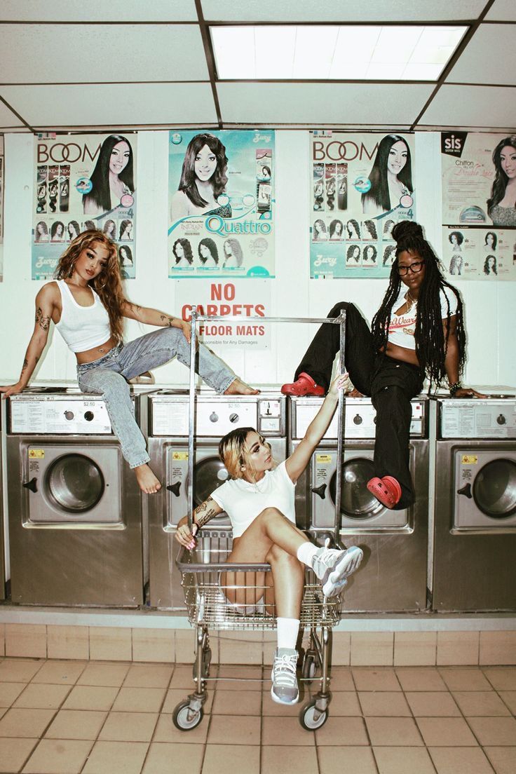 two women sitting on top of a shopping cart in front of washers and dryers