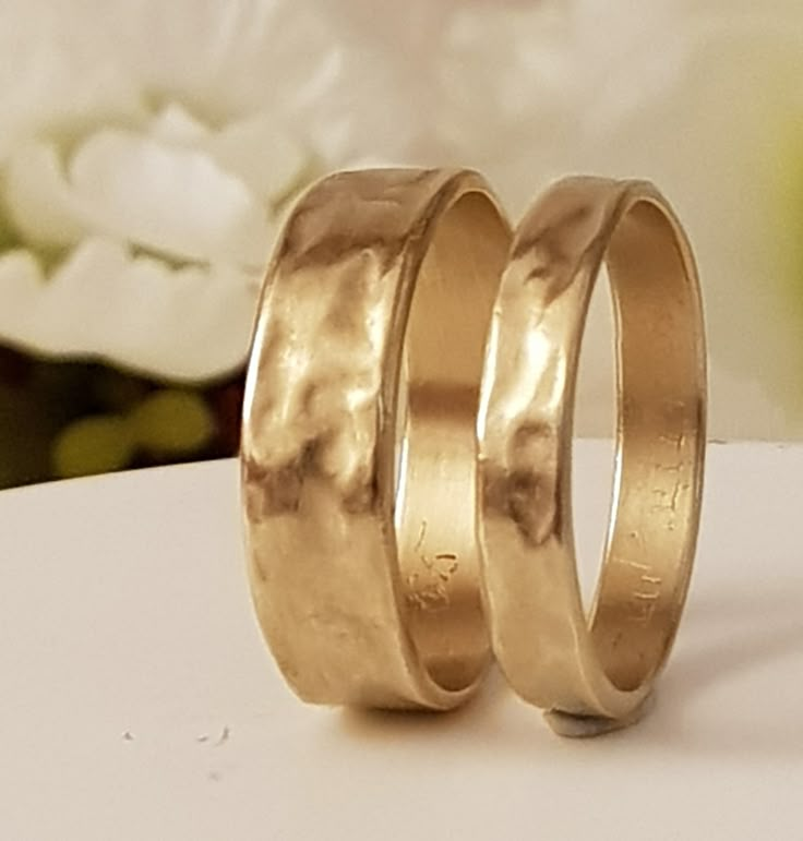 two gold wedding rings sitting next to each other on top of a white table with flowers in the background