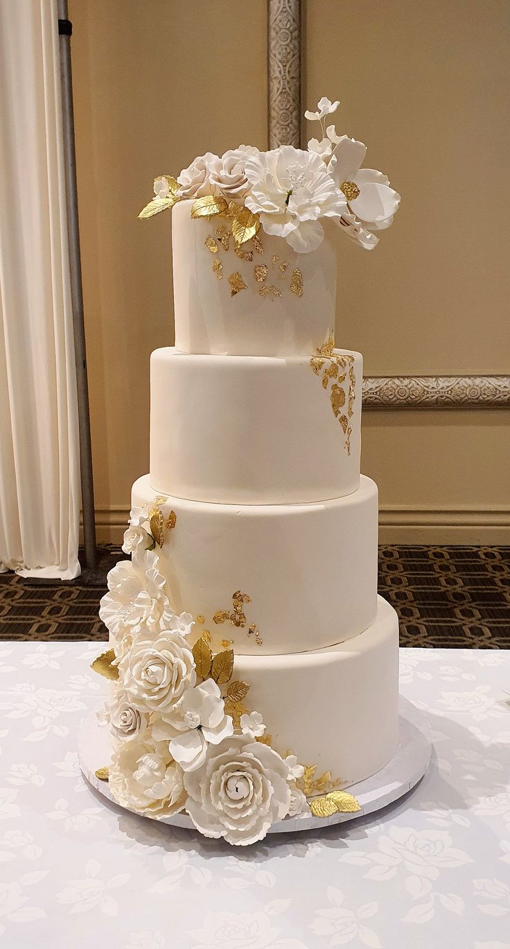 a three tiered wedding cake with white and gold flowers on the top, sitting on a table