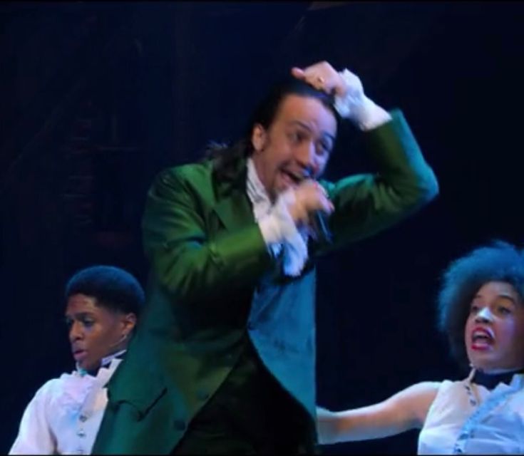 a man in a green suit and white shirt dancing with other men on stage behind him