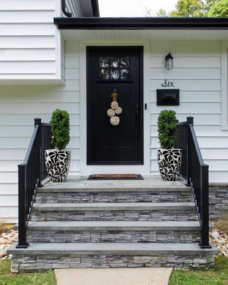 a black front door with two planters on the steps and a white house in the background