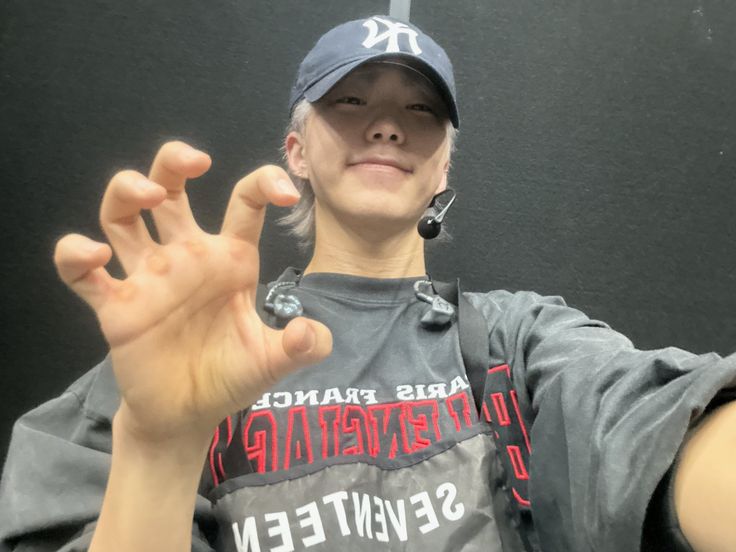 a woman wearing a baseball cap making the v sign with her hands while sitting in front of a black wall