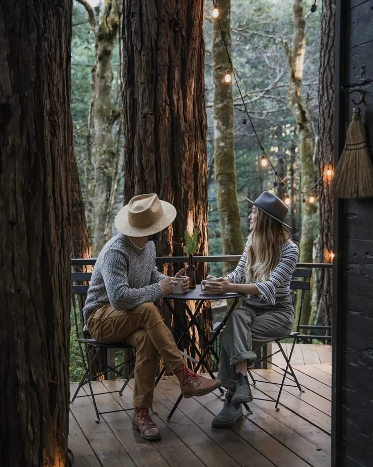 two people sitting at a table in the middle of a forest with lights strung from trees