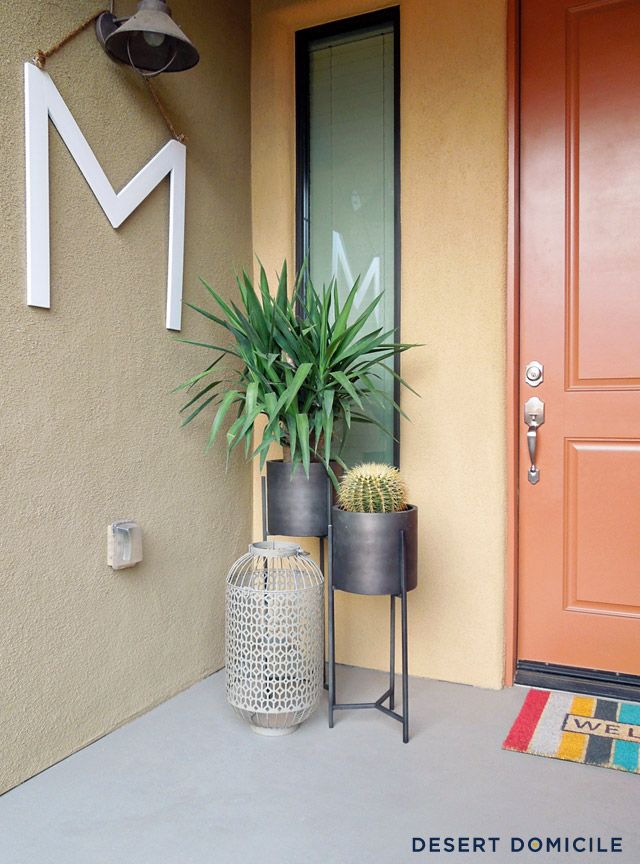 two potted plants sitting on the front porch next to a red door with an m sign above it