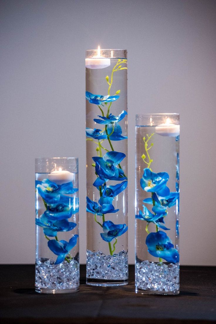 three tall glass vases with blue flowers and candles in them on a wooden table