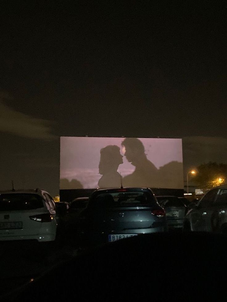 a couple kissing in front of a large screen at night with cars parked around them