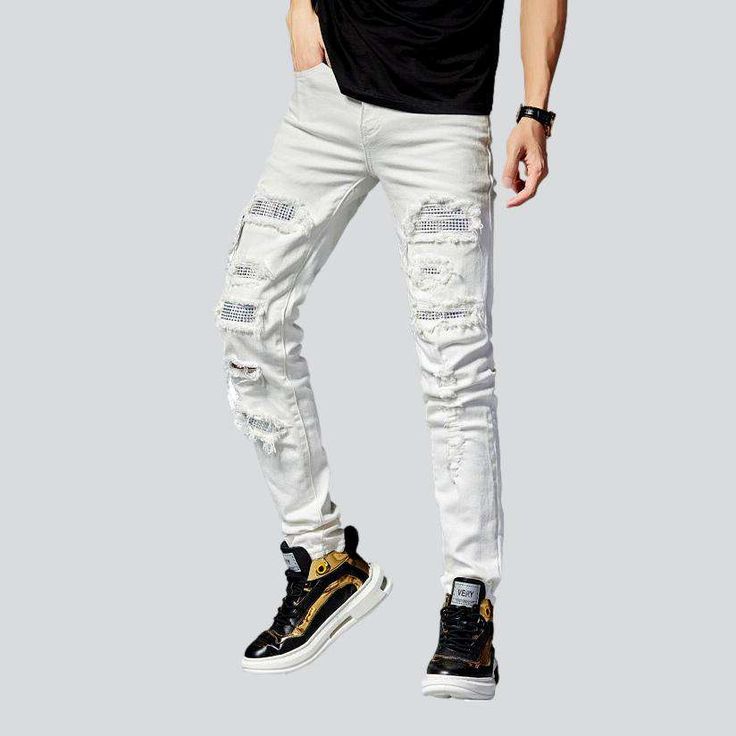 a man in black shirt and white ripped jeans standing on grey background with his hands in his pockets