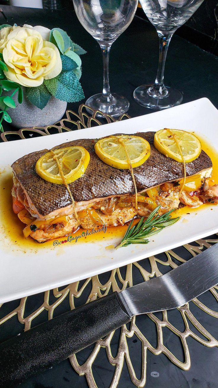 a white plate topped with fish covered in lemons and sauce next to two wine glasses