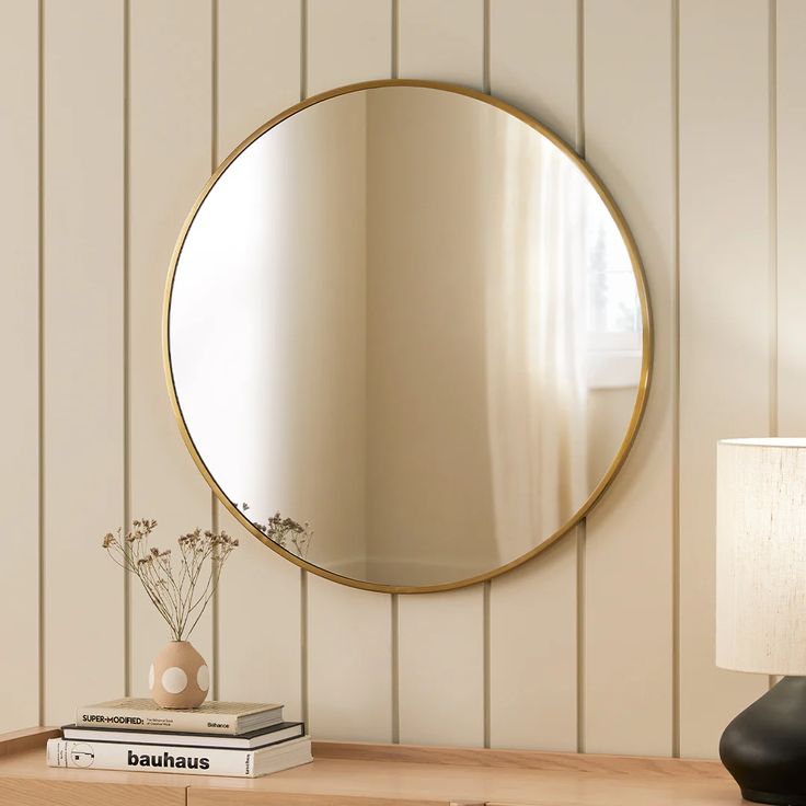 a round mirror sitting on top of a wooden table next to a lamp and book