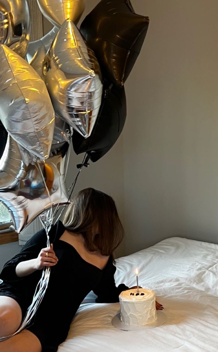 a woman is laying on her bed with balloons and a cake in front of her