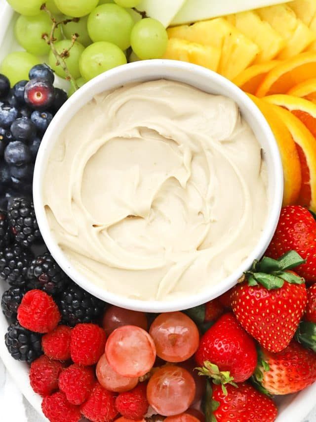 a white bowl filled with fruit and dip surrounded by grapes, apples, oranges, strawberries