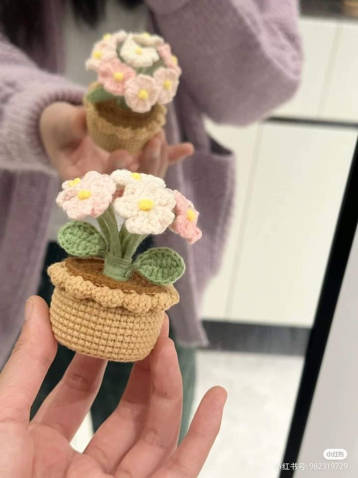 two small crocheted pots with flowers on them are held in front of a mirror