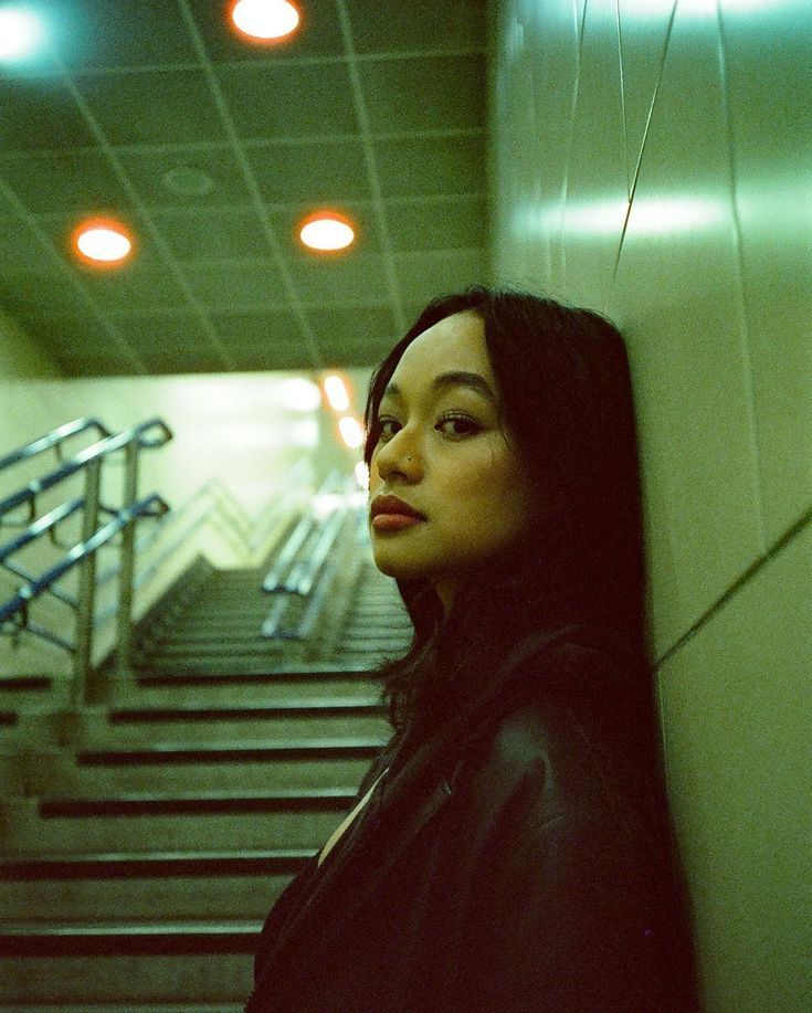 a woman leaning against a wall next to some stairs with lights on the ceiling above her