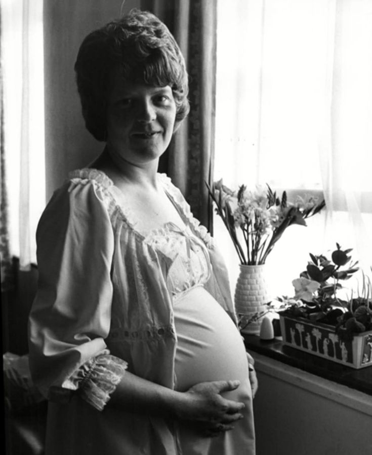 a pregnant woman standing in front of a window next to a vase with flowers on it