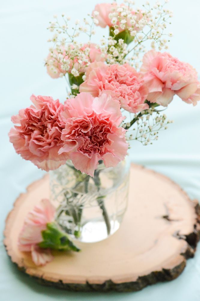 pink carnations and baby's breath in a glass vase on a wood slice