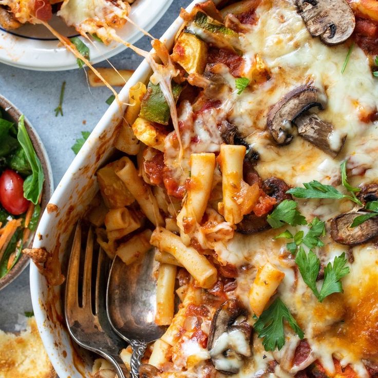 a casserole dish filled with pasta, mushrooms and spinach leaves next to a salad