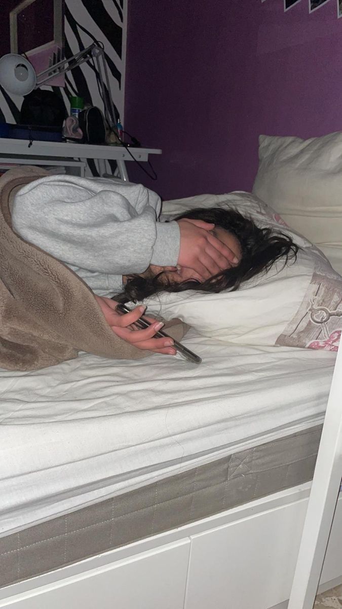 a woman laying on top of a white bed next to a purple wall and holding a cell phone