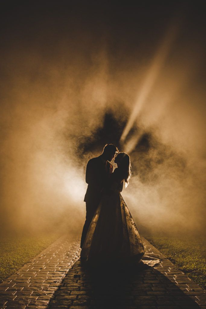 a bride and groom standing in the fog on their wedding day at night with light coming from behind them