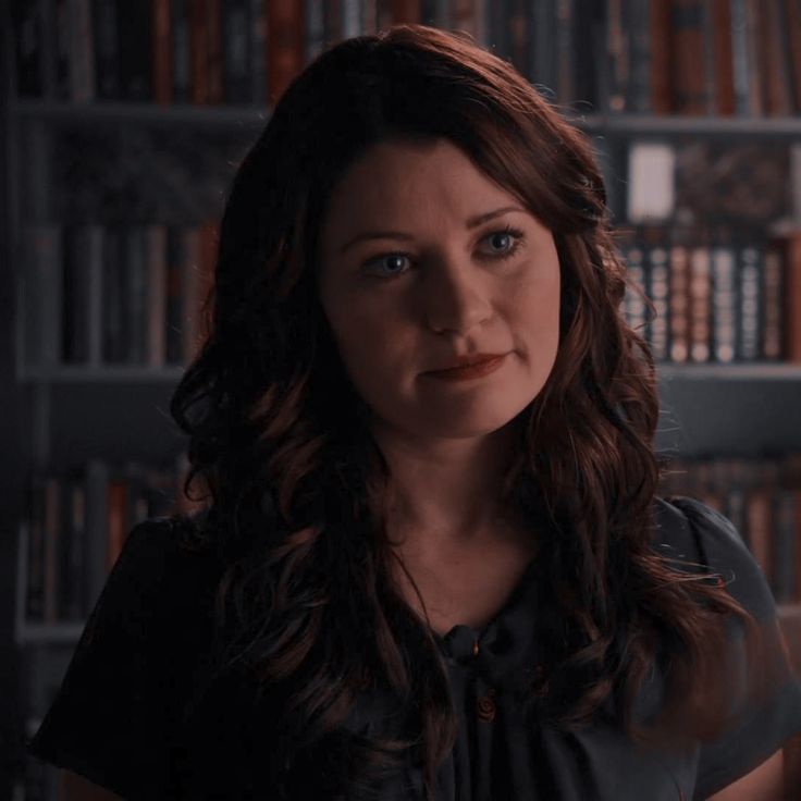 a woman with long hair standing in front of bookshelves and looking at the camera