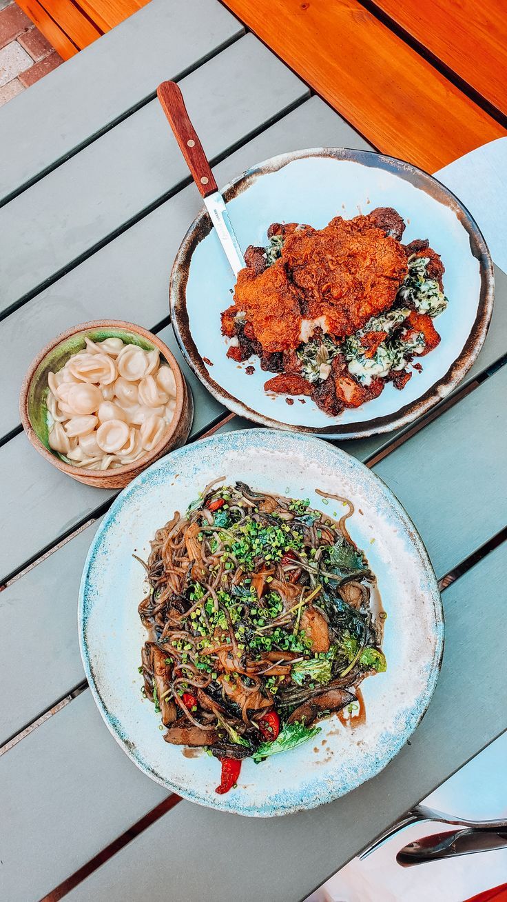 two plates of food sitting on top of a wooden table next to bowls of noodles