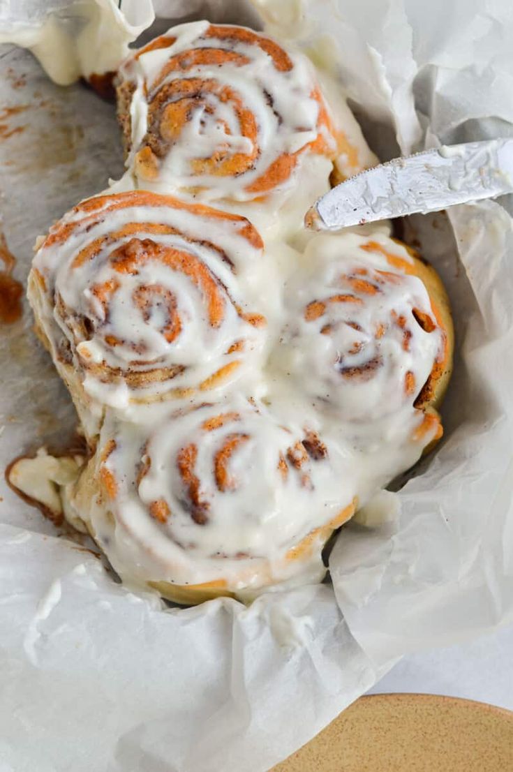 a cinnamon roll with icing sitting on top of a piece of wax paper next to a knife