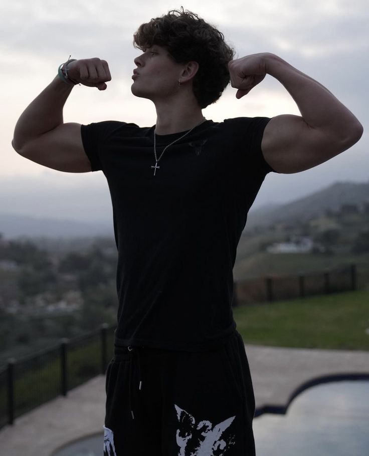 a young man flexing his muscles in front of a swimming pool and hills behind him