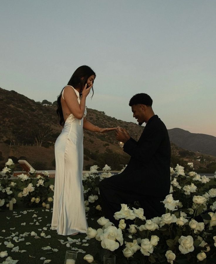 a man kneeling down next to a woman in a white dress holding a cell phone