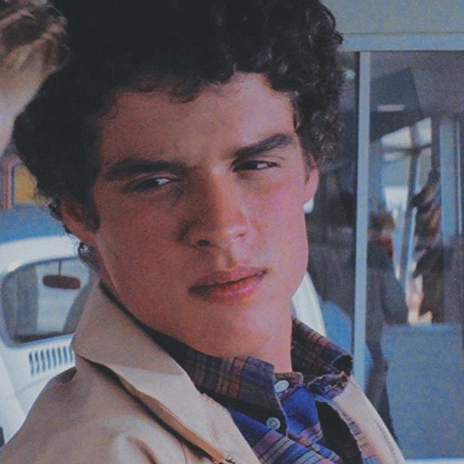 a young man with curly hair wearing a trench coat and looking at the camera while standing in front of a car