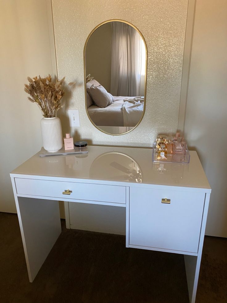 a white desk topped with a mirror and a vase filled with flowers next to it