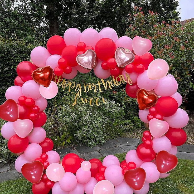 a heart - shaped balloon arch with balloons and hearts on it in front of some bushes