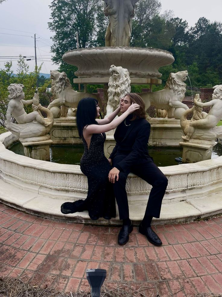 two people sitting on the edge of a fountain with their arms around each other's neck