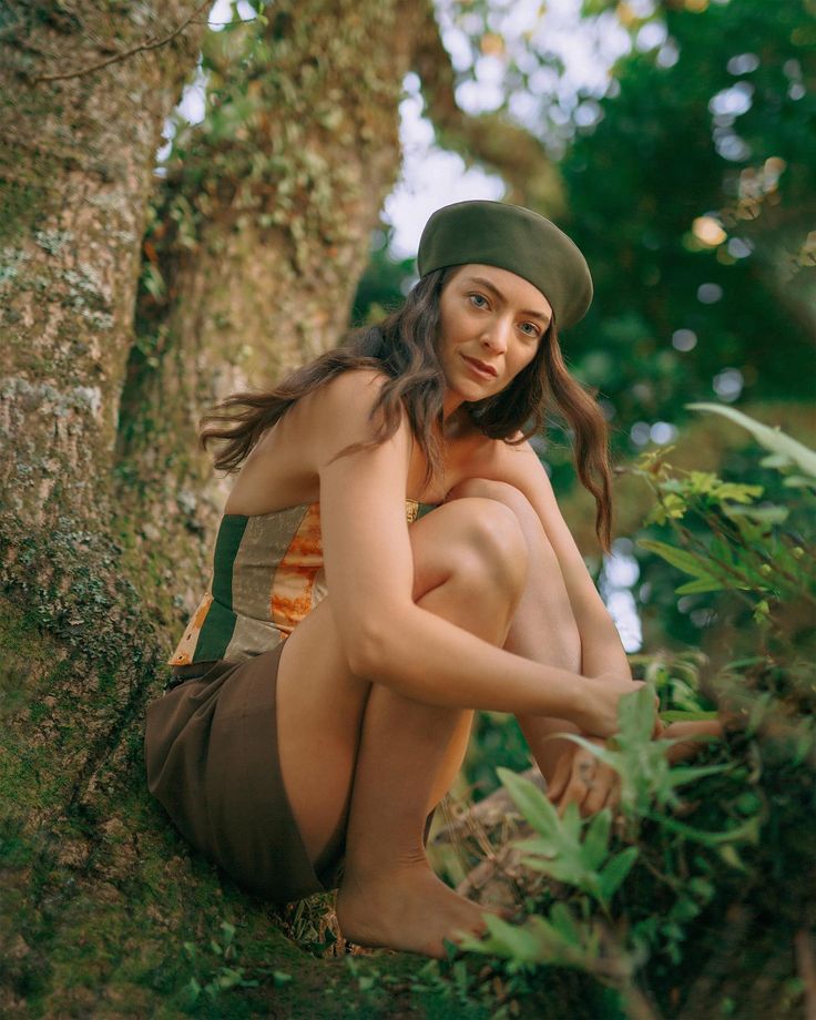 a woman sitting on the ground next to a tree wearing a green hat and brown skirt