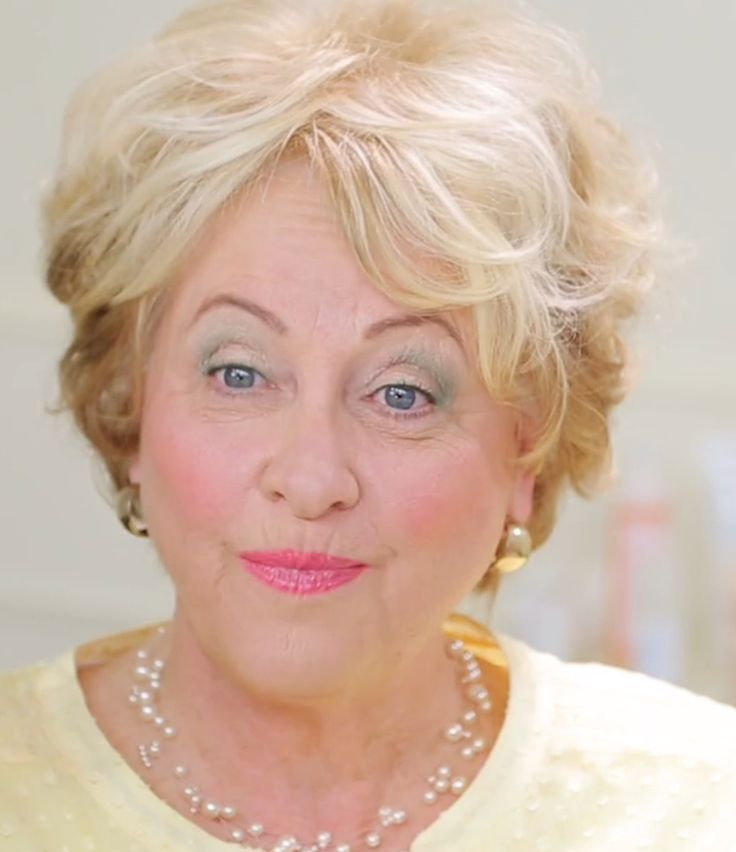 an older woman with blonde hair and pearls on her necklace looks at the camera while wearing a yellow sweater