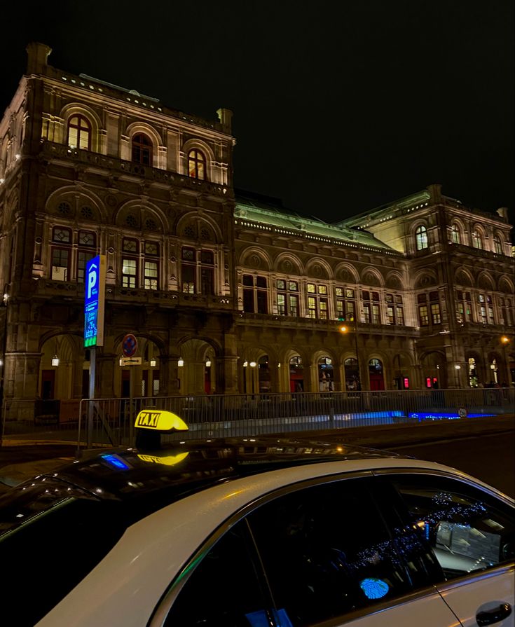 a white car parked in front of a large building at night with lights on it's windows