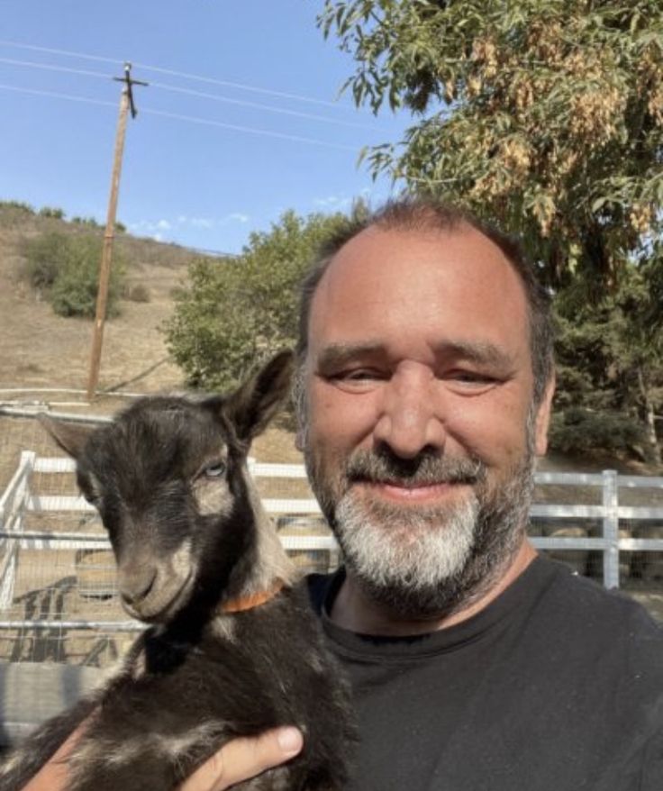 a man is holding a small goat in his arms and smiling at the camera while standing next to a fence