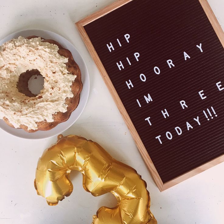 a doughnut and some gold foil balloons on a white table with a sign that says hip hooray i'm three today