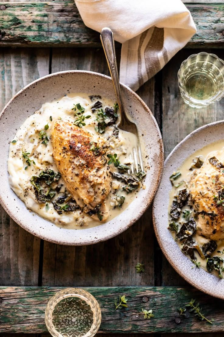 two plates with chicken and mashed potatoes covered in sauce on a wooden table next to a glass of water