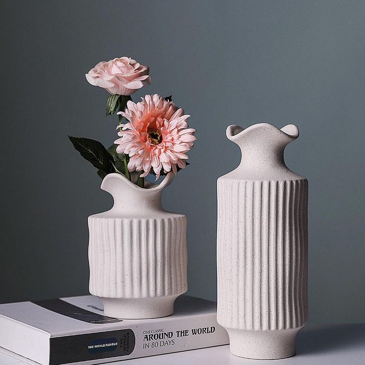 two white vases with pink flowers in them on top of a stack of books