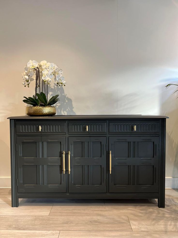 a vase with flowers sitting on top of a black buffet sideboard against a white wall