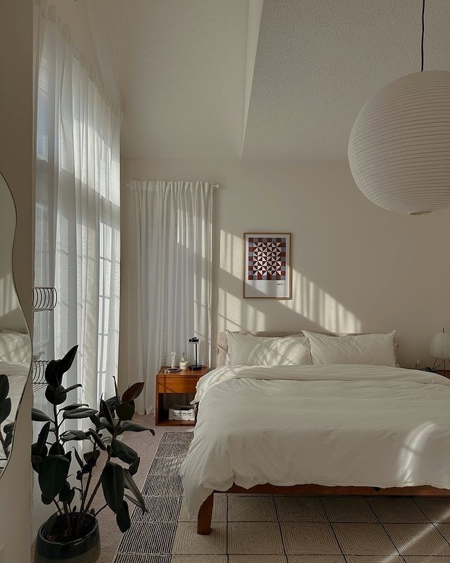 a white bed sitting in a bedroom next to a tall window filled with curtains and potted plants